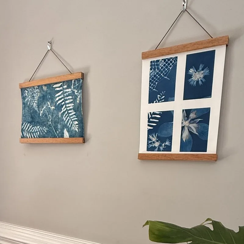 Two finished cyanotype prints hanging on a wall in wooden magnetic frames, a ferns print and a four-square botanical grid